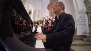 A pianist in formal attire plays a grand piano in a brightly lit church, with a choir and conductor in the background.