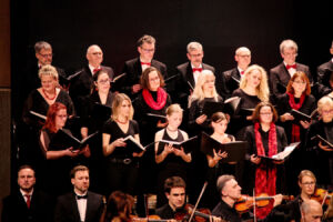 A choir and orchestra performing on stage, with choir members dressed in black holding songbooks and musicians playing string instruments in front.