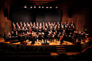 A large choir and orchestra are performing on stage in a concert hall, illuminated by stage lights.