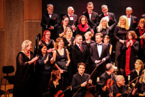 A choir and orchestra on stage, dressed formally, pose together with smiles and applause, suggesting the end of a performance.