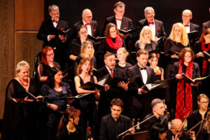 A choir of men and women dressed in formal black and red attire performs on stage. They hold open music sheets and sing in harmony, with musicians visible in front of them.
