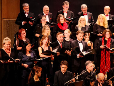 A choir of men and women dressed in formal black and red attire performs on stage. They hold open music sheets and sing in harmony, with musicians visible in front of them.