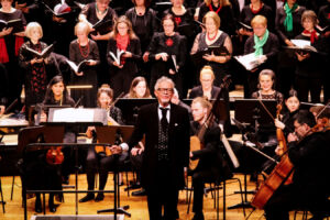 A choir and orchestra perform on stage with a conductor in front, wearing formal attire. The choir, dressed in black with red and green accents, holds music sheets. The orchestra members play various instruments, creating a harmonious scene.