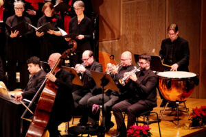 Musicians performing in an orchestra, featuring brass and percussion instruments. Choir members are singing in the background.