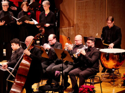 Musicians performing in an orchestra, featuring brass and percussion instruments. Choir members are singing in the background.