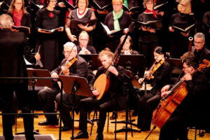 An orchestra and choir perform on stage, musicians play various string instruments, and choir members hold sheet music while singing in the background.