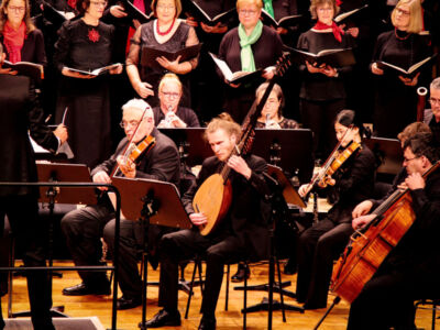 An orchestra and choir perform on stage, musicians play various string instruments, and choir members hold sheet music while singing in the background.