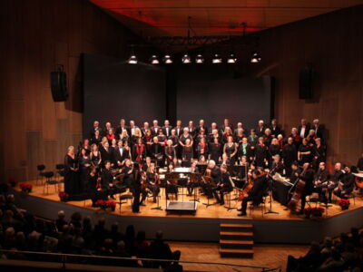 An orchestra and choir perform on a dimly lit concert hall stage, with musicians playing strings and woodwinds, and vocalists standing in rows behind them. An audience is seated in the foreground.