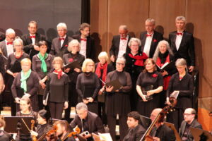 A choir in formal attire stands on stage, some holding sheet music. Musicians are seated with their instruments in the foreground.