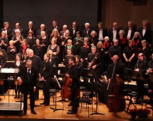 An orchestra and choir take a bow on stage, with musicians holding various instruments and vocalists standing in the background, all dressed in formal attire.