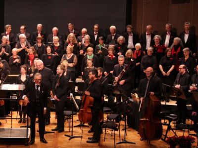 An orchestra and choir take a bow on stage, with musicians holding various instruments and vocalists standing in the background, all dressed in formal attire.