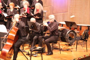 Musicians perform in an orchestra with string, brass, and percussion instruments. A choir stands behind them on stage, singing in unison.
