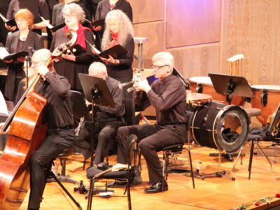 Musicians perform in an orchestra with string, brass, and percussion instruments. A choir stands behind them on stage, singing in unison.