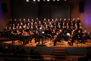 A choir and orchestra perform on a well-lit stage in a concert hall, featuring a conductor directing musicians and singers dressed in black formal attire.