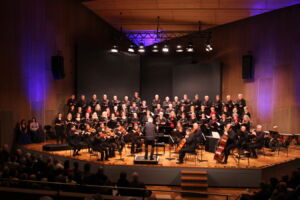 A choir and orchestra perform on a large stage in a concert hall with a conductor leading.