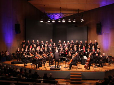 A choir and orchestra perform on a large stage in a concert hall with a conductor leading.