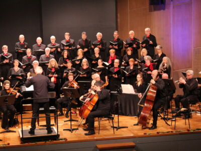 A choir and orchestra perform on stage, with the conductor leading. The choir members stand in rows, holding music sheets, while the musicians are seated with various instruments. The setting appears formal, with a focus on musical performance.