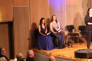 Two women seated on a stage in formal gowns, holding sheet music.
