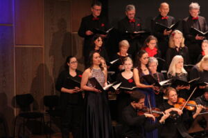 A choir of men and women, dressed in formal black attire, perform on stage, holding sheet music. A conductor and violinists are positioned in front, adding orchestral accompaniment.