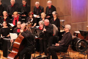 A choir and musicians perform on stage, dressed in black, with a focus on a double bass player and a flutist. The choir members, also in black with red accents, are seated behind them holding sheet music.