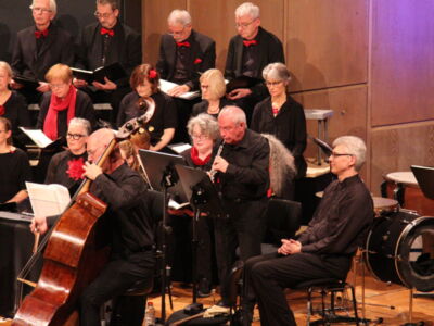 A choir and musicians perform on stage, dressed in black, with a focus on a double bass player and a flutist. The choir members, also in black with red accents, are seated behind them holding sheet music.