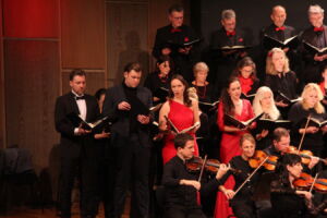 A choir and orchestra perform on stage, with singers dressed in black and red, and musicians playing string instruments.