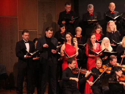 A choir and orchestra perform on stage, with singers dressed in black and red, and musicians playing string instruments.