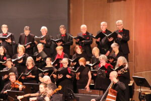 A choir and orchestra perform on stage, with singers dressed in black and musicians playing instruments like violins and cellos.