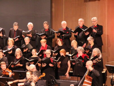 A choir and orchestra perform on stage, with singers dressed in black and musicians playing instruments like violins and cellos.