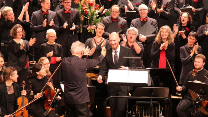 A conductor faces an orchestra and choir on stage, receiving applause and cheers. The musicians are dressed in formal black attire with red accents. The atmosphere is celebratory and joyful.
