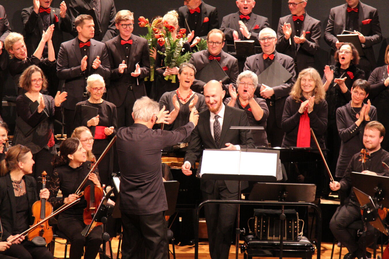A conductor faces an orchestra and choir on stage, receiving applause and cheers. The musicians are dressed in formal black attire with red accents. The atmosphere is celebratory and joyful.