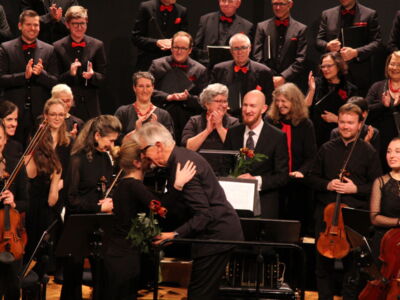 A choir and orchestra on stage applaud a conductor and a musician sharing a congratulatory hug, with several musicians holding their instruments and wearing formal black attire.