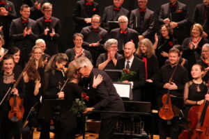 A conductor embraces a musician on stage after a performance, surrounded by an orchestra and choir members dressed in black.