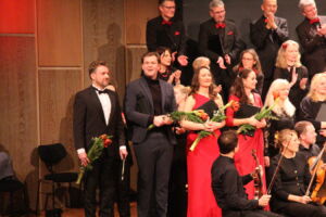 Performers on stage holding flowers, standing beside musicians in formal attire, with an audience applauding in the background.