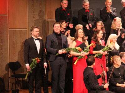 Performers on stage holding flowers, standing beside musicians in formal attire, with an audience applauding in the background.