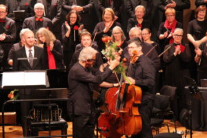 A cellist receives flowers on stage amid applause from a standing choir ensemble, all dressed in black with red accents.