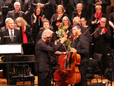 A cellist receives flowers on stage amid applause from a standing choir ensemble, all dressed in black with red accents.