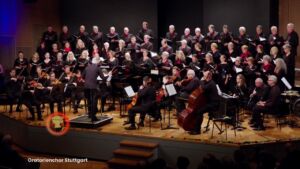 A large choir and orchestra perform on stage with a conductor leading them. The choir members are standing in rows, while the orchestra sits in front with string and wind instruments. The stage is well-lit, and the audience is visible in the foreground.