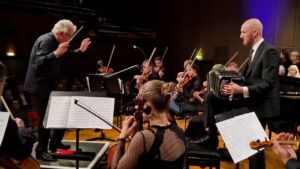 An orchestra performs on stage with a conductor and musicians, including a man playing an accordion. The ensemble is focused on their music, creating a dynamic and engaged atmosphere.