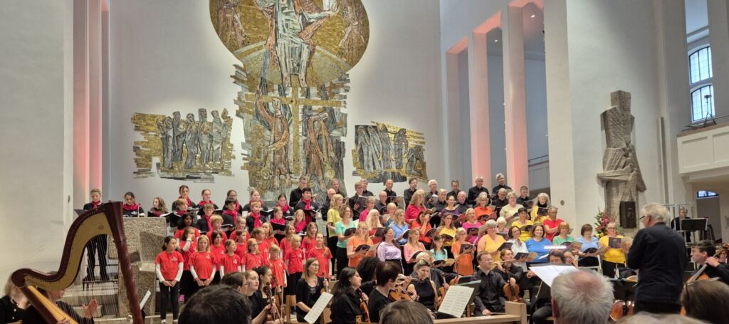 A large choir wearing colorful attire performs on a well-lit stage in a spacious hall, accompanied by an orchestra. The backdrop features a prominent mural and tall pillars, with an audience visible in the foreground.
