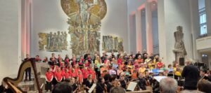 A large choir wearing colorful attire performs on a well-lit stage in a spacious hall, accompanied by an orchestra. The backdrop features a prominent mural and tall pillars, with an audience visible in the foreground.