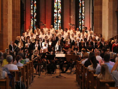 A large choir with musicians performs in a church with stained glass windows, while an audience watches from wooden pews.