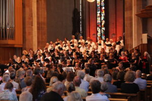 A large choir performs in a church with stained glass windows and a pipe organ visible. An audience is seated in pews, watching the performance.