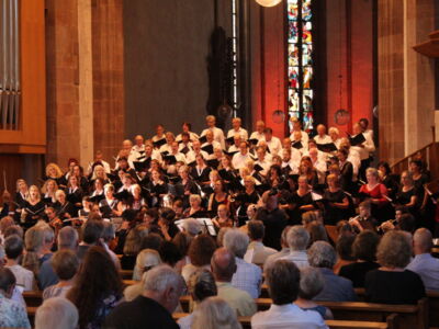 A large choir performs in a church with stained glass windows and a pipe organ visible. An audience is seated in pews, watching the performance.