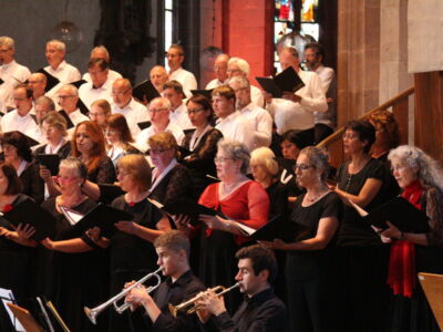 A choir dressed in black and white performs in a church, with members holding songbooks. Two musicians play a trumpet and a clarinet in front of the choir. Stained glass windows are visible in the background.