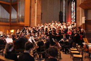 An orchestra and choir perform in a large, ornate church with stained glass windows, surrounded by wooden pews and organ pipes.