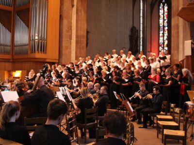 An orchestra and choir perform in a large, ornate church with stained glass windows, surrounded by wooden pews and organ pipes.
