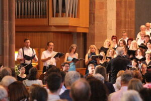 A choir performs in a church setting, led by a conductor. The singers hold sheet music, and an audience watches the performance. The backdrop includes an organ and wooden paneling.