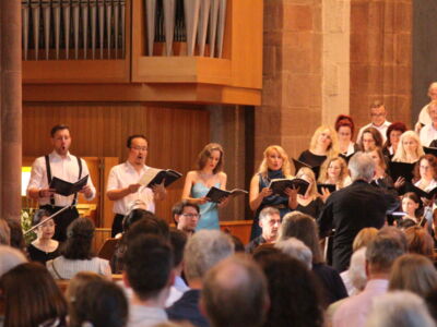 A choir performs in a church setting, led by a conductor. The singers hold sheet music, and an audience watches the performance. The backdrop includes an organ and wooden paneling.