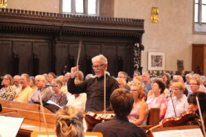 A conductor energetically leads an orchestra in front of a seated audience inside a warmly lit church or hall, with individuals attentively watching the performance.